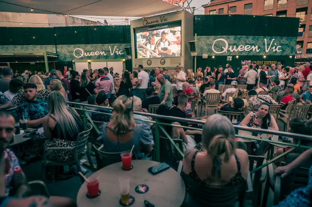 Queen Vic's packed terrace at dusk in Lloret de Mar, giant screen showing the match under festoon lights